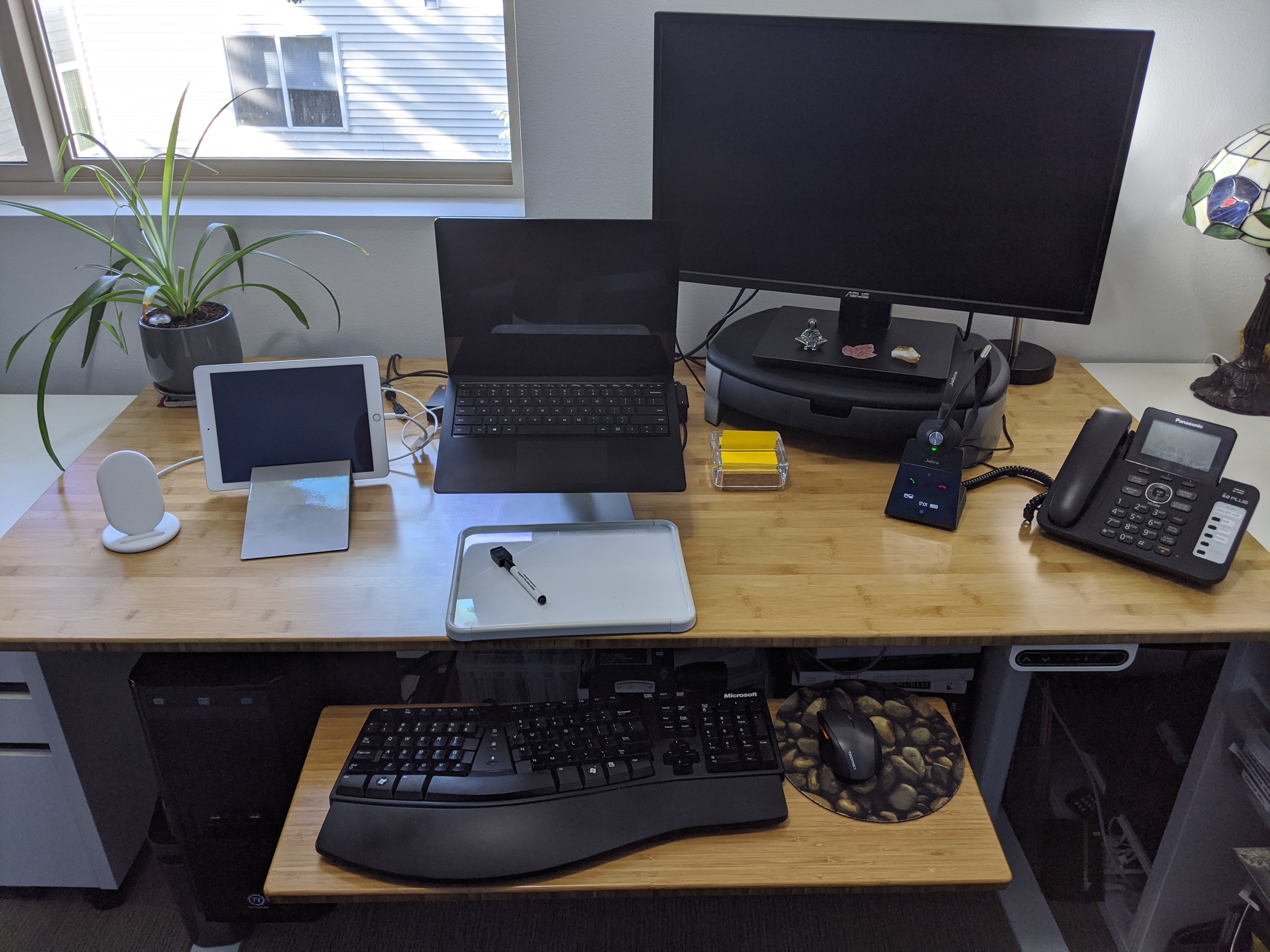Bamboo desk with laptop, monitor, keyboard, iPad, phone and headset. Window in background.